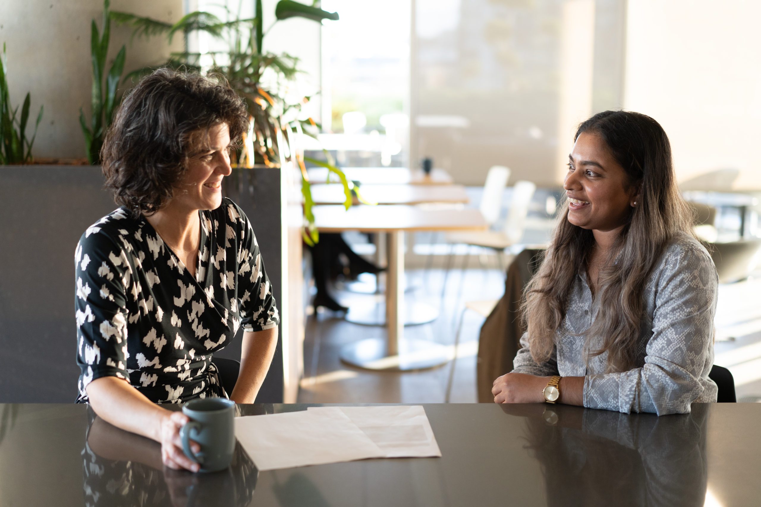 Two BCI employees, one holding a mug of coffee, sitting at a counter in the lunchroom having a conversation. Two BCI employees, one holding a mug of coffee, sitting at a counter in the lunchroom having a conversation.